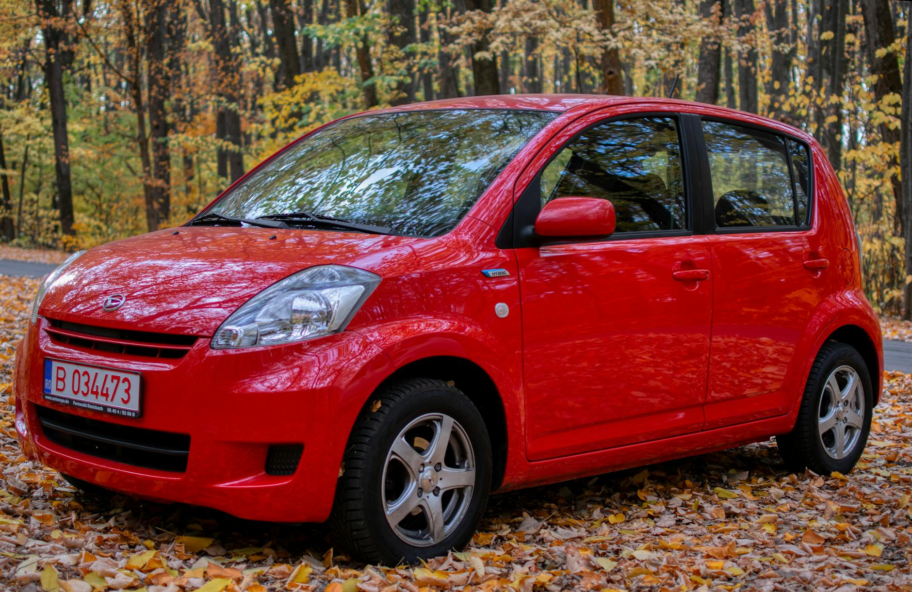 A parked red Daihatsu hatchback on a leaf-covered road in fall.