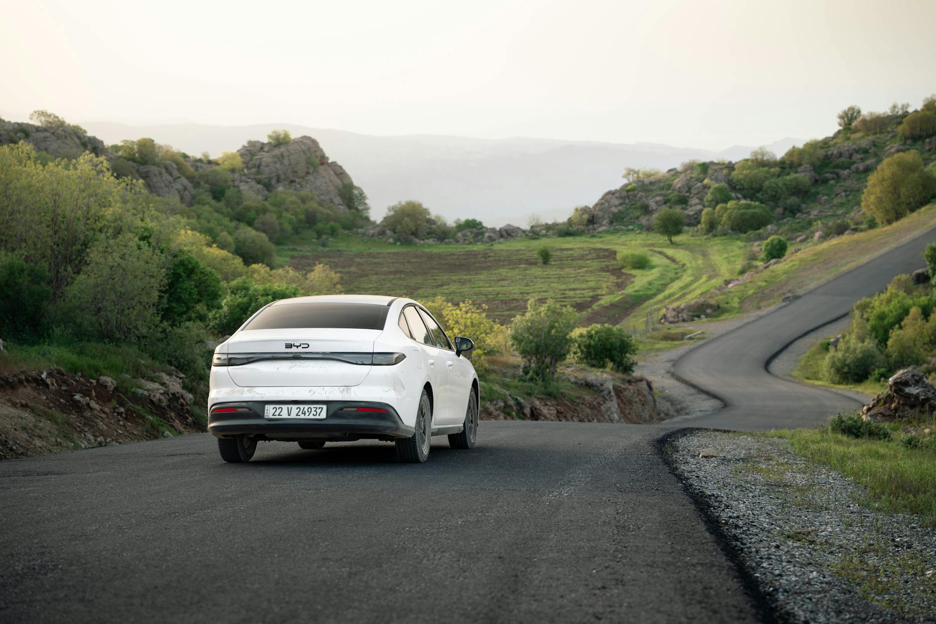 A white car drives on a winding road through scenic hills in Shaqlawa, Iraq during daylight.