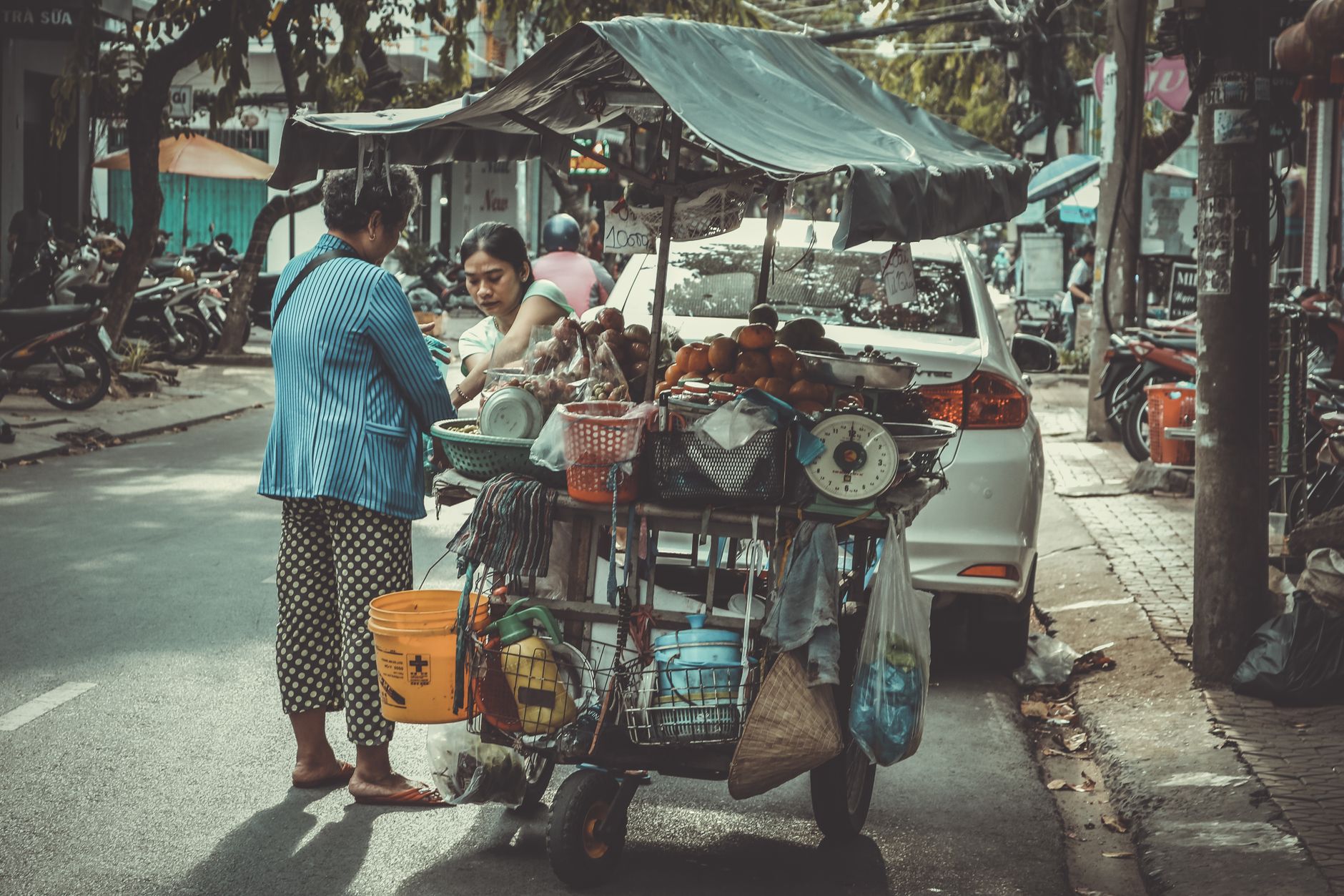Two female vendors selling produce from a mobile cart on a busy street in Vietnam.