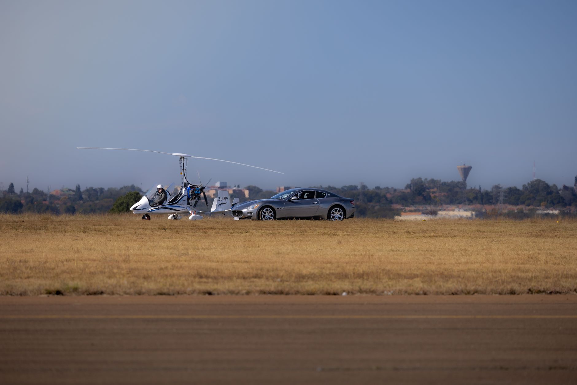 A sleek gyrocopter and a sports car side by side on a sunny airfield.