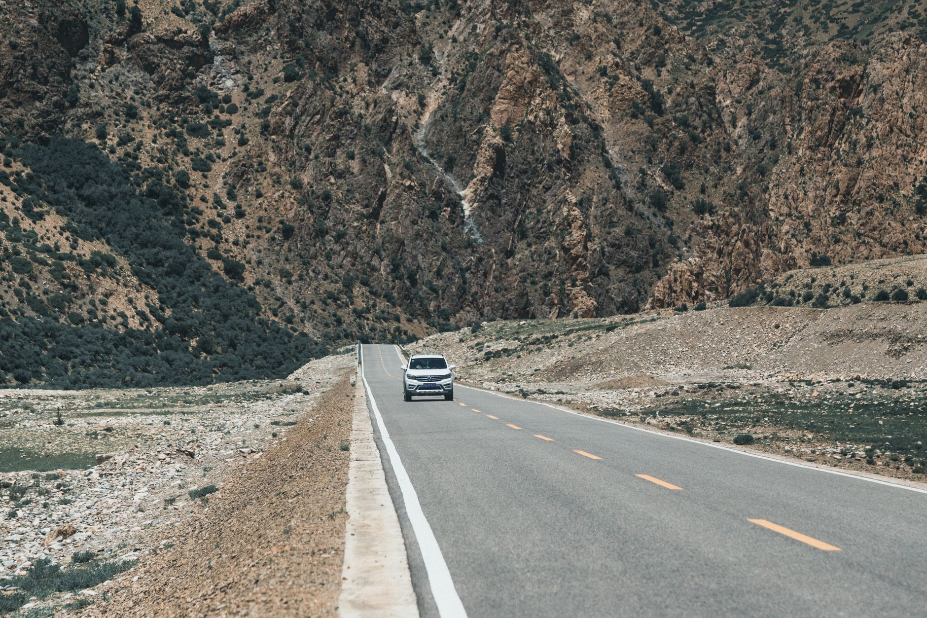 A white SUV travels on a deserted road through the rugged mountains of Tibet, under a clear sky.