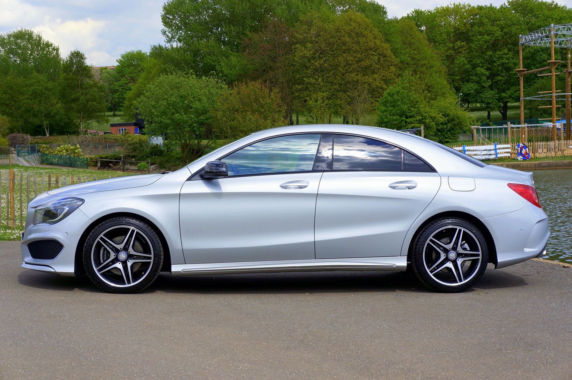 Side view of a silver luxury car parked on a road beside a scenic lake surrounded by trees.
