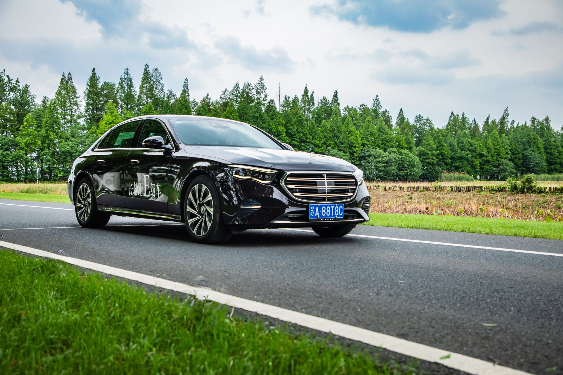 A sleek black luxury sedan drives along a scenic country road with lush greenery.