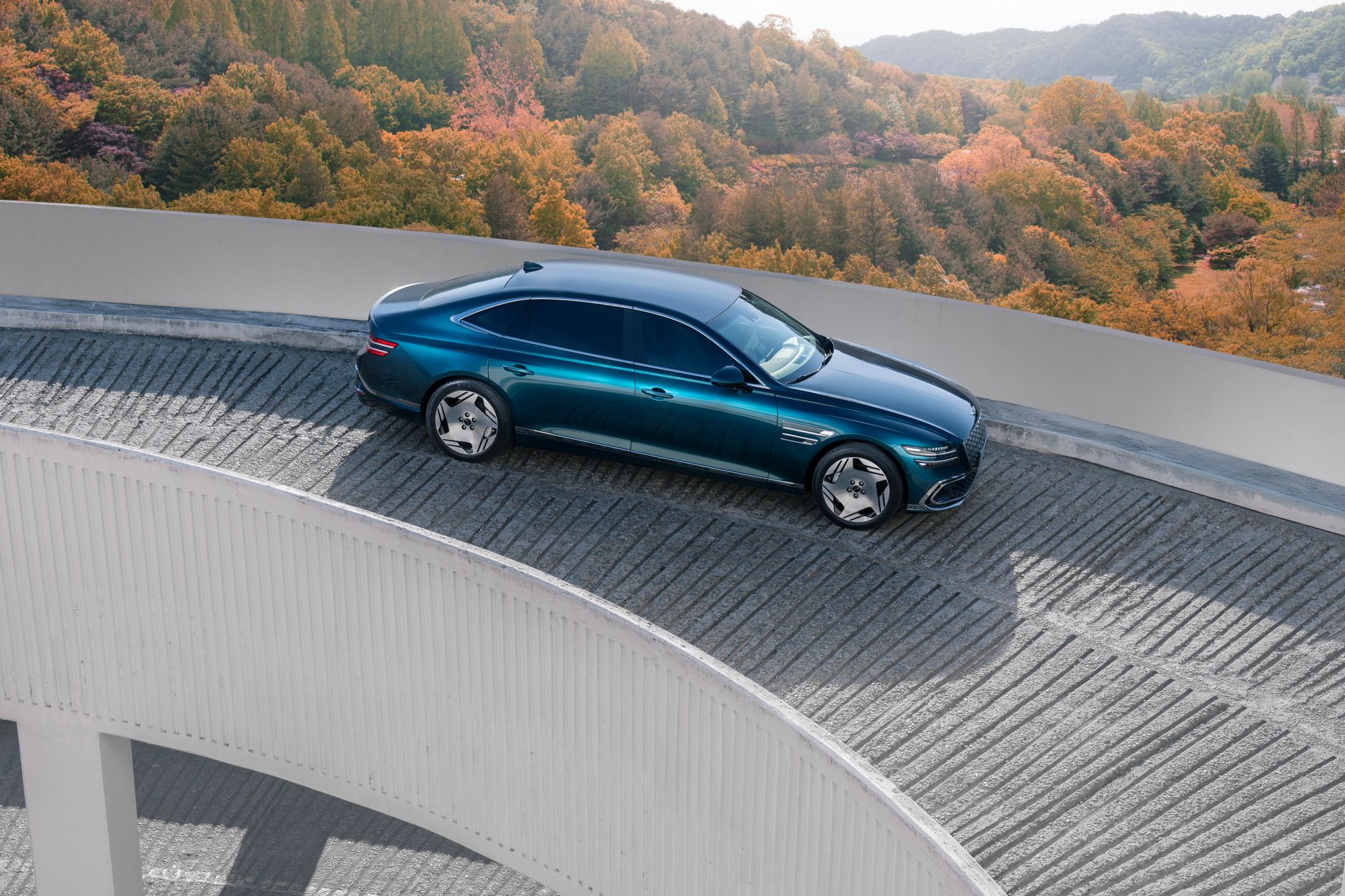 A blue luxury electric car on a curved road with autumn foliage in the background.