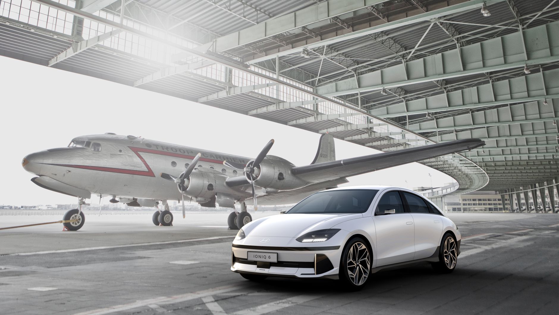 Sleek white electric car parked near a vintage aircraft in an open hangar.