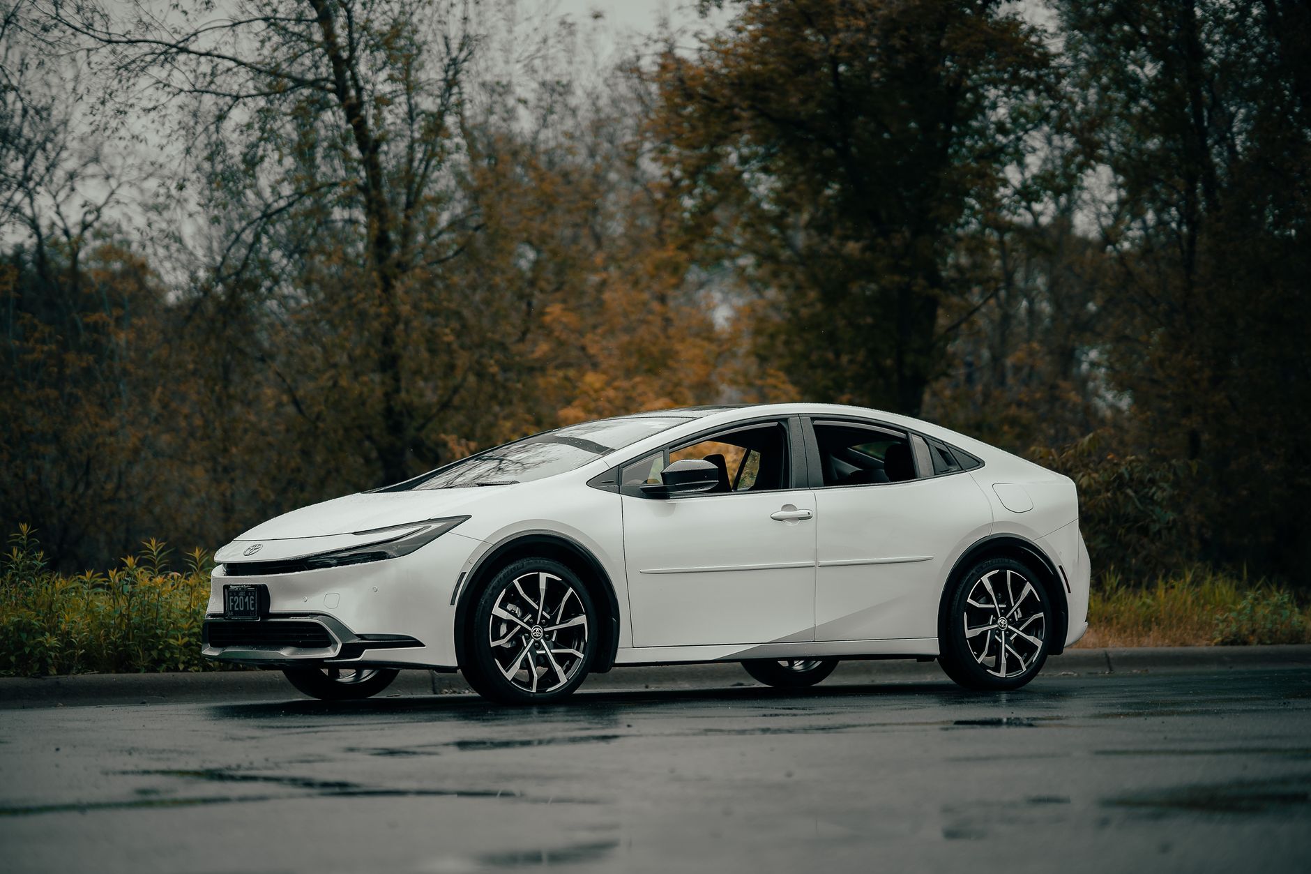 Side view of a modern white sedan car on a wet road surrounded by autumn trees.