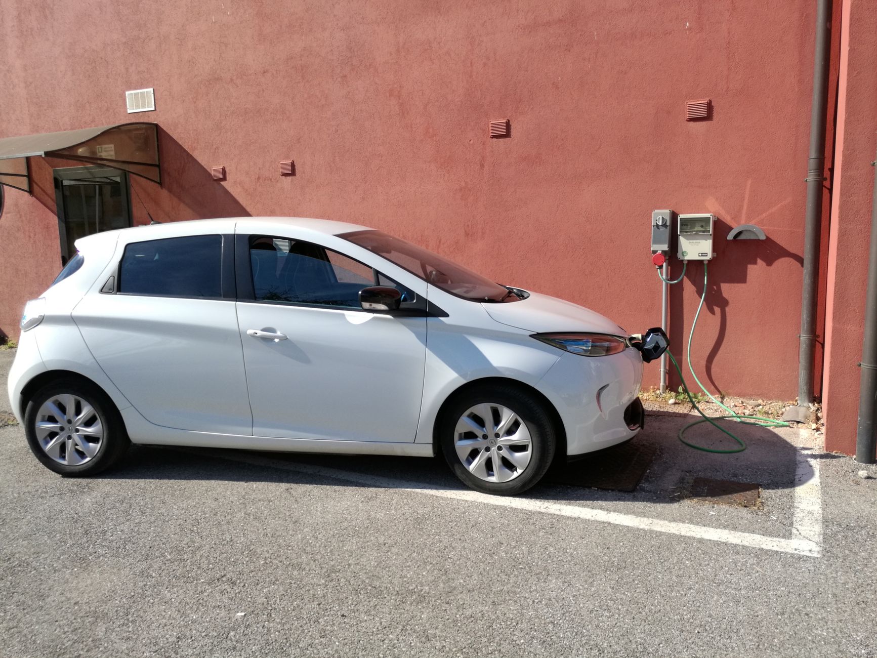 White electric vehicle charging at station against a red wall in San Bartolomeo di Breda di Piave, Italy.