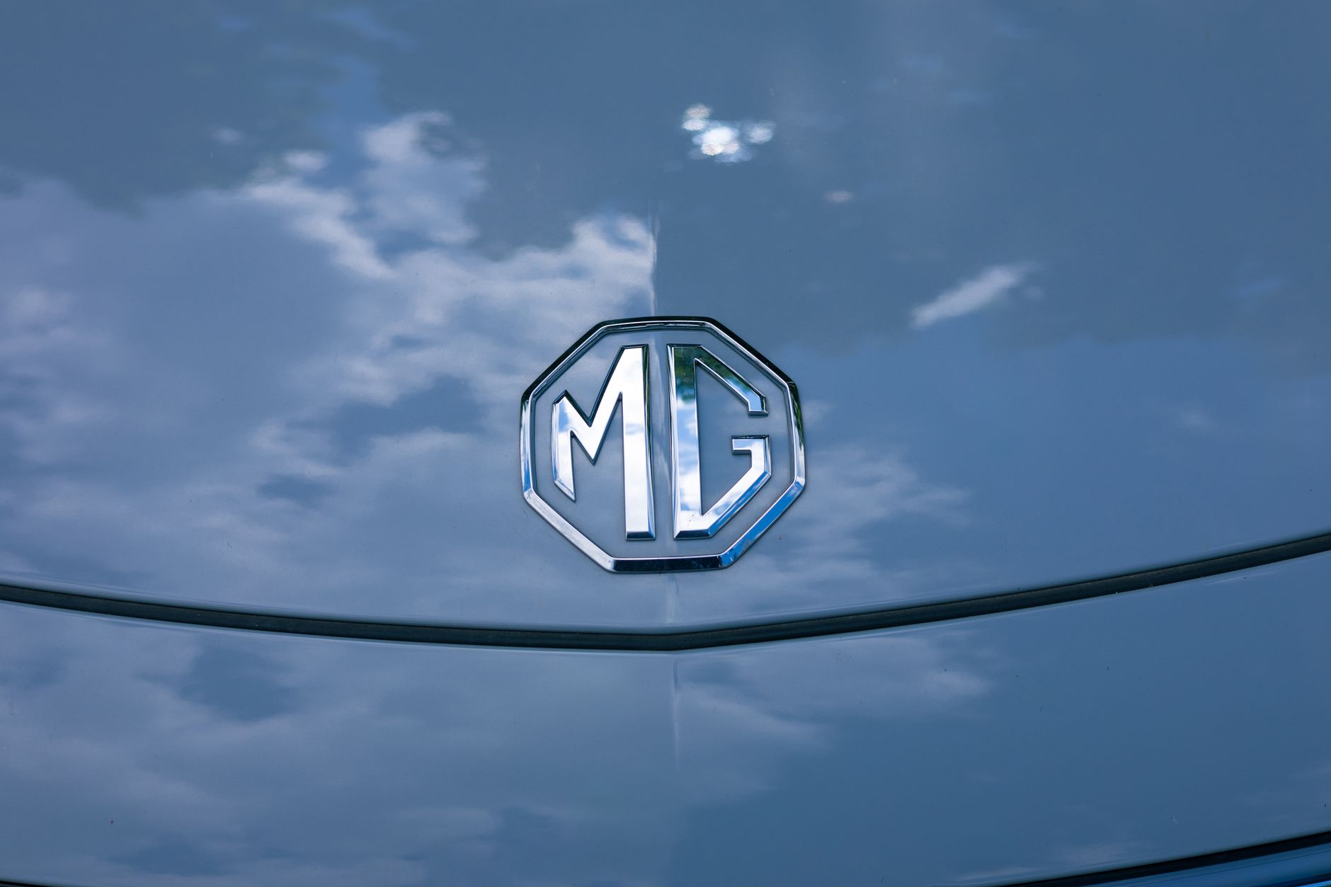 Vibrant MG logo on a car hood with striking sky reflection, symbolizing elegance.