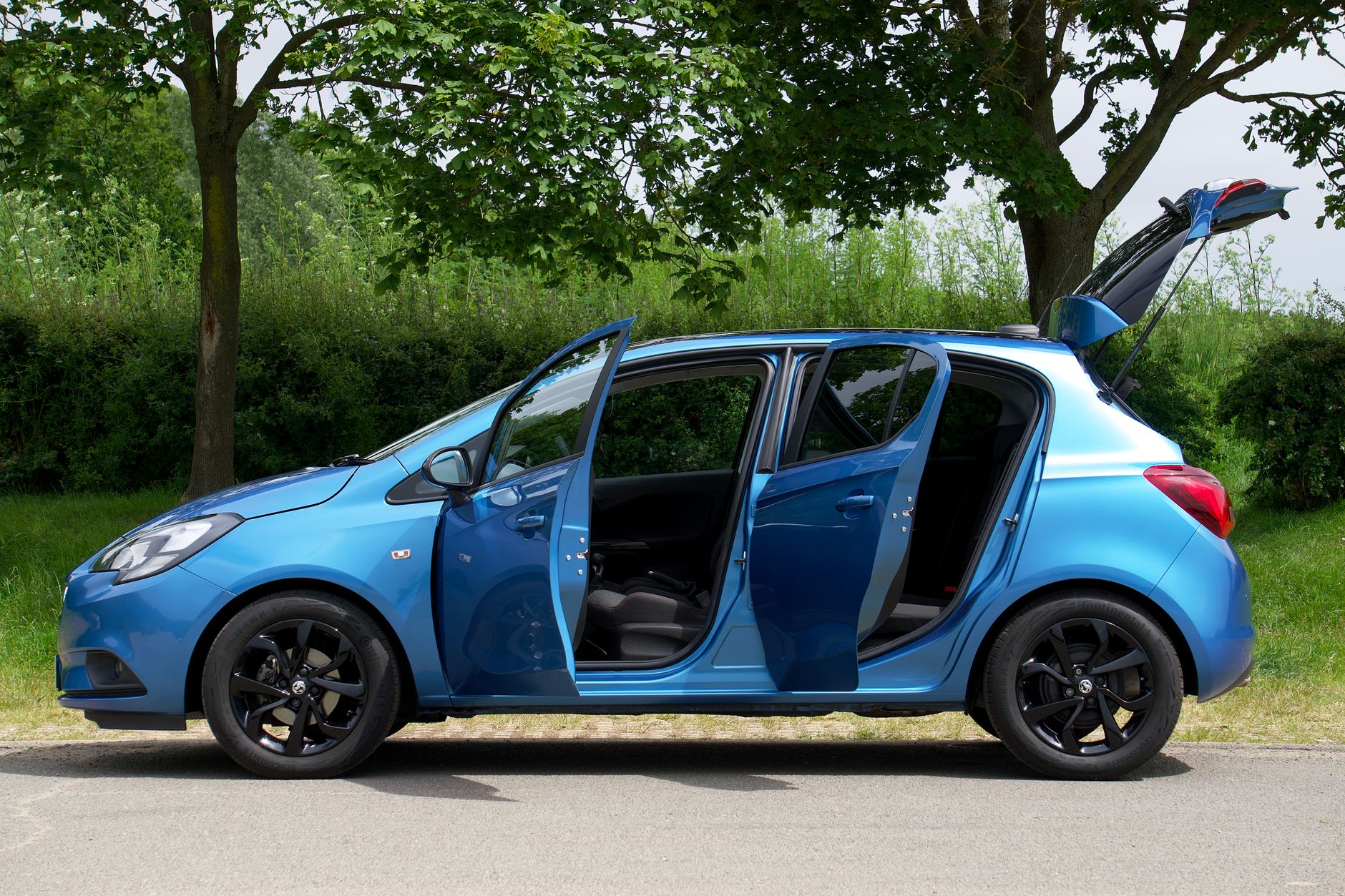 A vibrant blue hatchback with open doors parked on a road amidst lush greenery.