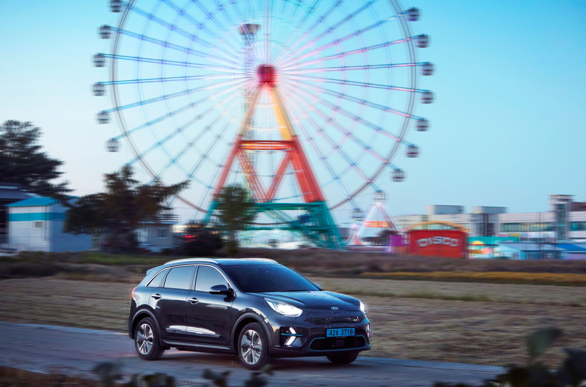 Electric SUV passes brightly lit Ferris wheel at amusement park.